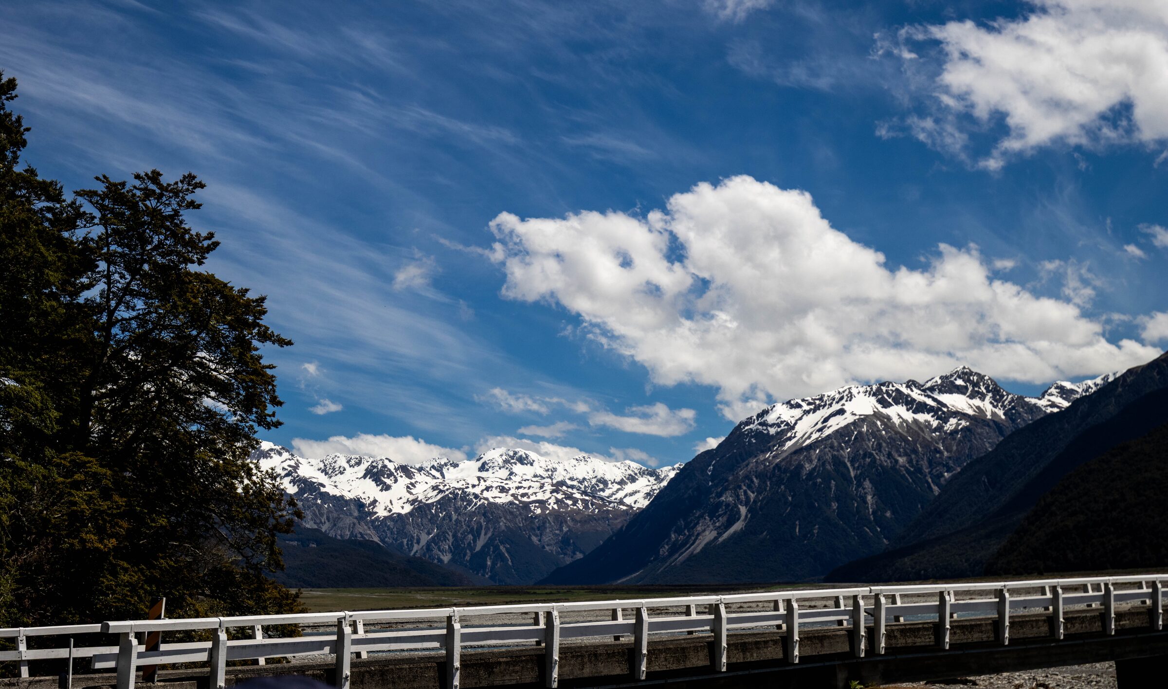 Snow-capped mountains above a braided river valley, with a tree and bridge railing in the foreground
